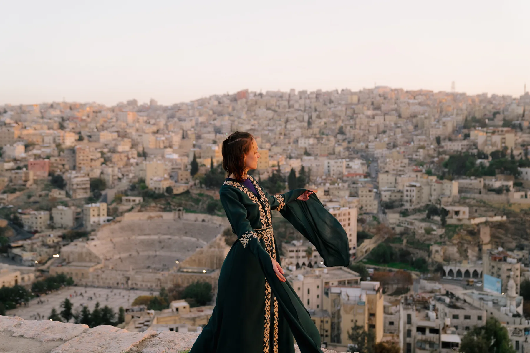 A traveller on the hillside above Amman at golden hour, the Roman theatre and the city behind her